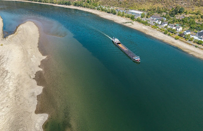 A cargo barge traveling along a wide river beside a sandy bank